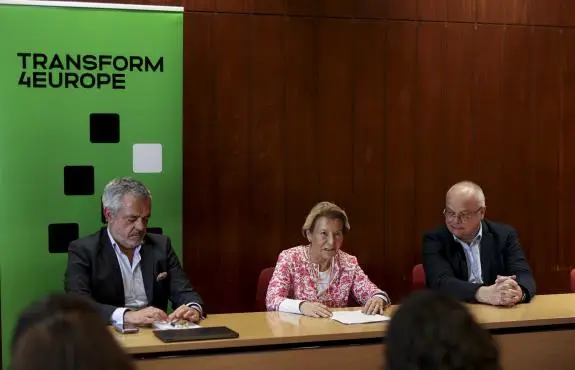 Three people sit at a table in front of a green Transform 4 Europe banner, engaged in a discussion or meeting. The woman in the center appears to be speaking, while the two men listen attentively.