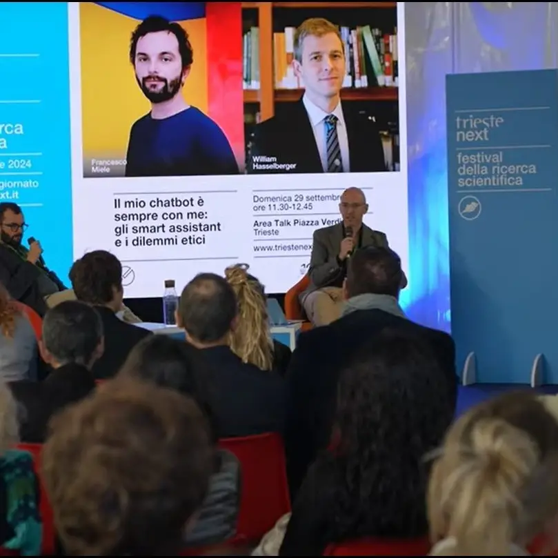 A speaker addresses an audience at a science festival. Behind him, a large screen displays headshots and names of Francesco Miele and William Hasselberger, along with event details in Italian. Blue festival banners are visible.