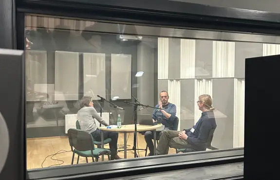 Three people are sitting around a table with microphones in a recording studio, seen through a glass window. They appear to be engaged in conversation or a podcast. Acoustic panels are on the walls.