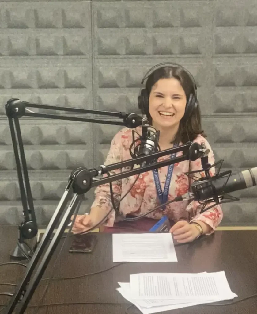 A woman wearing headphones and a floral shirt sits at a table with microphones, papers, and a phone in a soundproof recording studio, smiling at the camera.