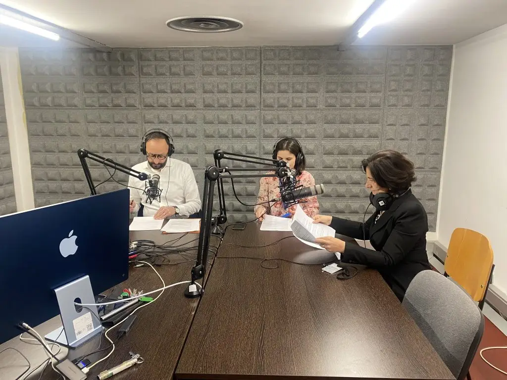 Three people sit at a table in a soundproofed recording studio, wearing headphones and speaking into microphones. Papers and a computer are on the table, suggesting they are recording a podcast or radio show.