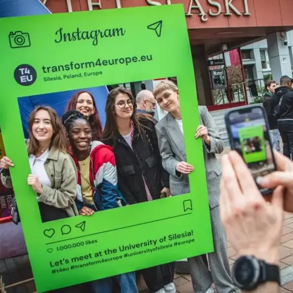 Five smiling young adults pose together outdoors, holding a large green Instagram-style photo frame promoting Transform4Europe at the University of Silesia, while someone takes their photo with a smartphone.