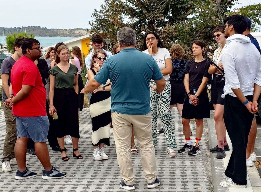 A group of people stands outdoors in a semi-circle, listening to a man in a blue shirt speaking. Trees and a building are visible in the background. Most people appear attentive, and the weather looks clear.