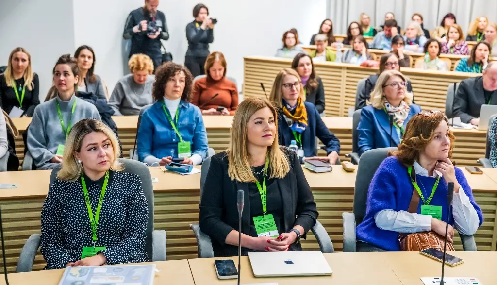 A group of women, many wearing green lanyards, sit attentively in a bright conference room. Several have laptops open on the desks in front of them, and more people are seated in the background.