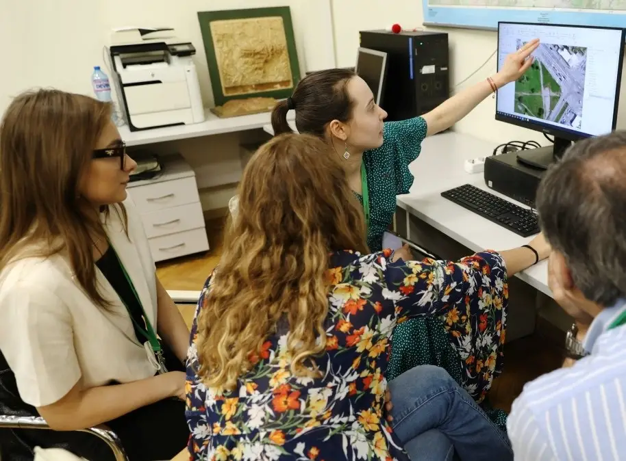 Four people sit around a desk in an office, closely looking at a computer monitor displaying a map or aerial photo. One woman is pointing at the screen while explaining something to the group.