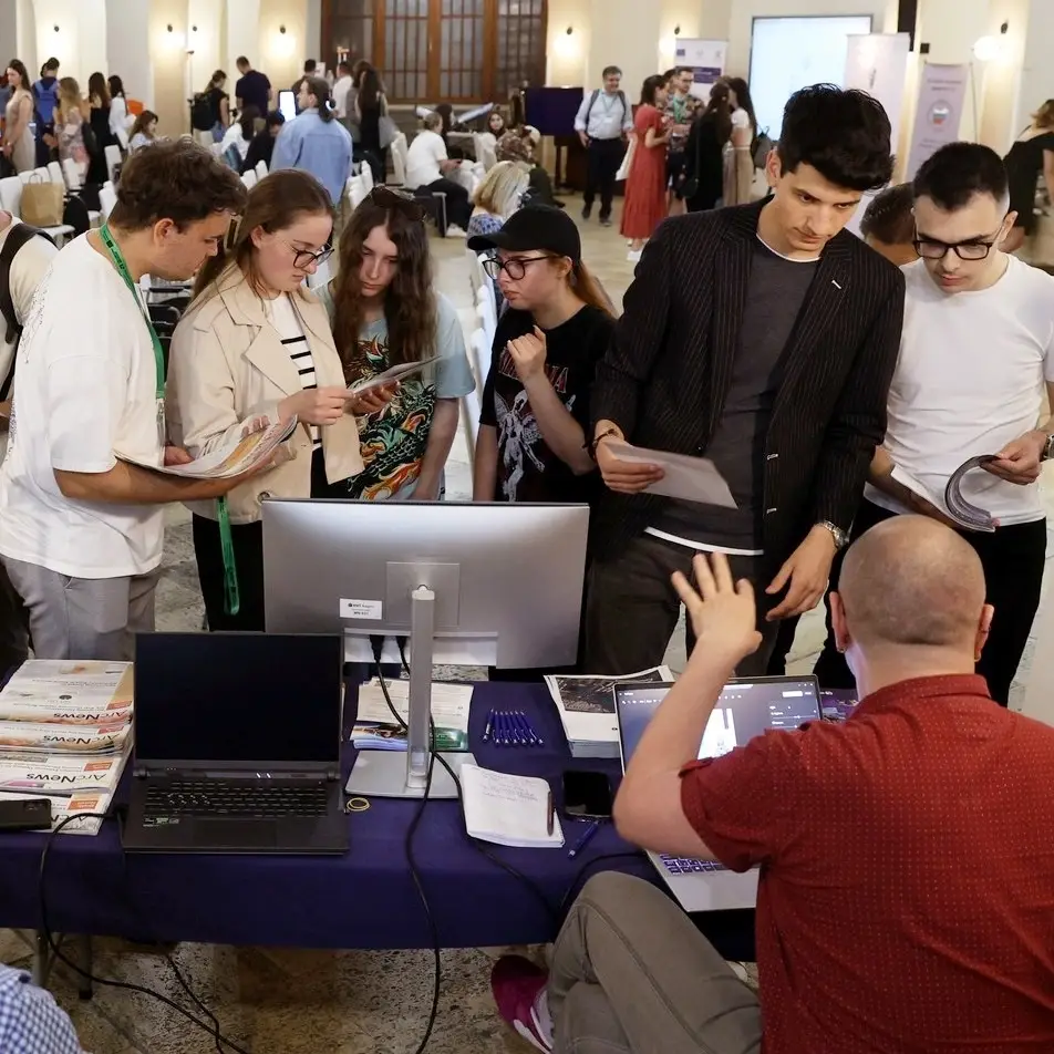 A group of young people stand around a table with computers and papers at an indoor event, engaging with a person seated who is gesturing and explaining something to them.