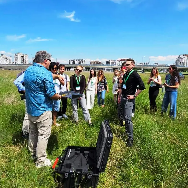 A group of people stand in a grassy field listening to a man holding a folder. Some wear lanyards, and buildings are visible in the background under a blue sky. An open black equipment case sits on the grass.