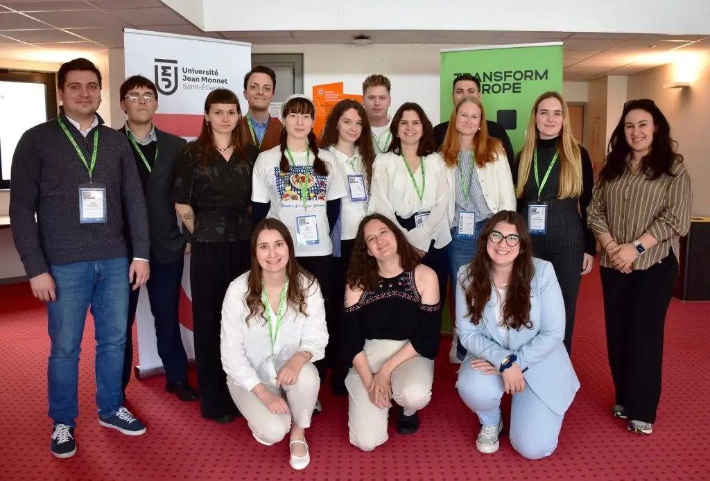 A group of thirteen smiling young adults and one older adult pose indoors, wearing name badges and green lanyards. Behind them are banners for “Université Jean Monnet” and “Transform Europe.”.