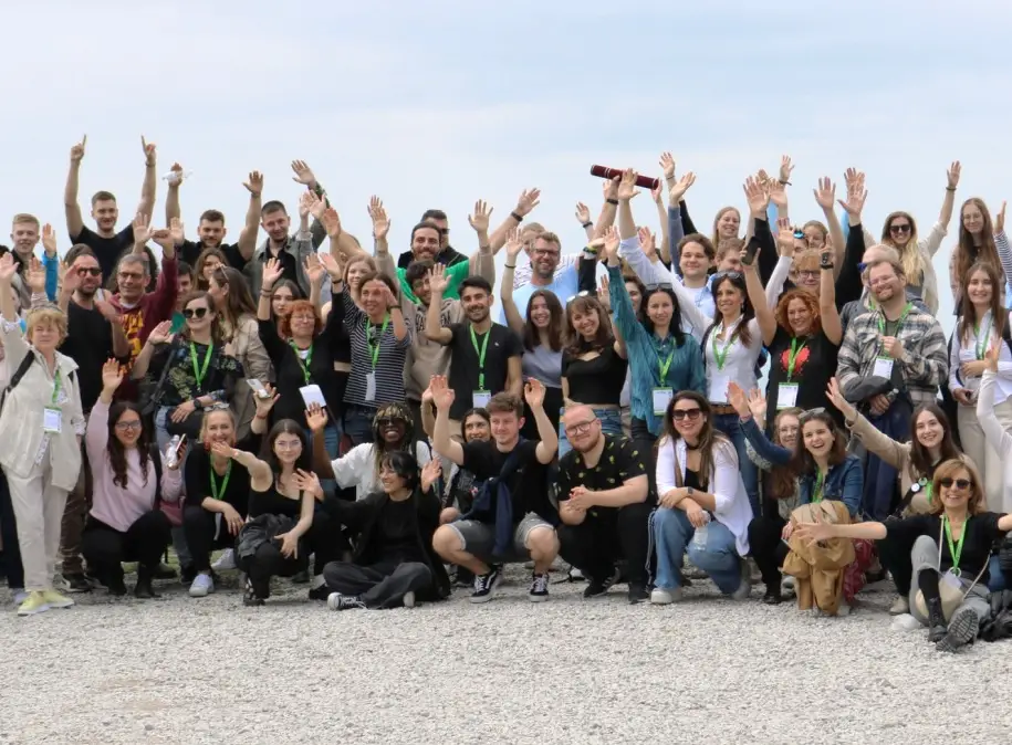 A large group of people outdoors, smiling and waving at the camera. Most are standing, some are sitting in front, and many wear name tags and lanyards. The sky is overcast and the ground is gravel.