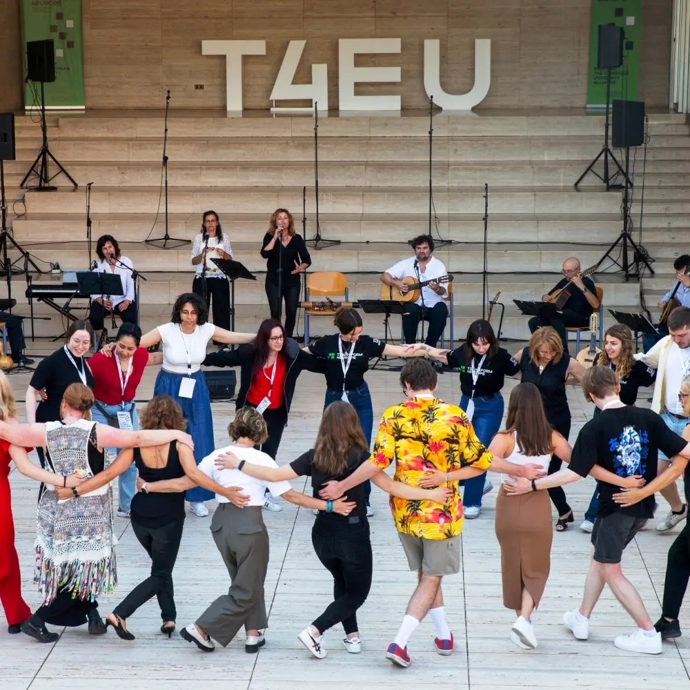 A group of people stands in a circle with arms around each other, dancing outdoors in front of a stage with live musicians and T4EU displayed above them. The scene is lively and communal.