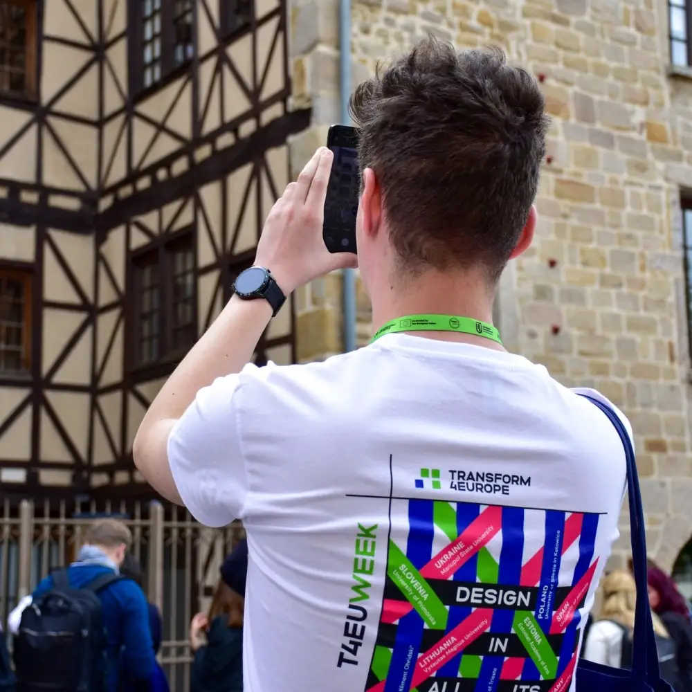 A person with short brown hair wearing a white Transform 4 Europe T-shirt takes a photo of a half-timbered building using a smartphone; a green lanyard and black watch are visible.