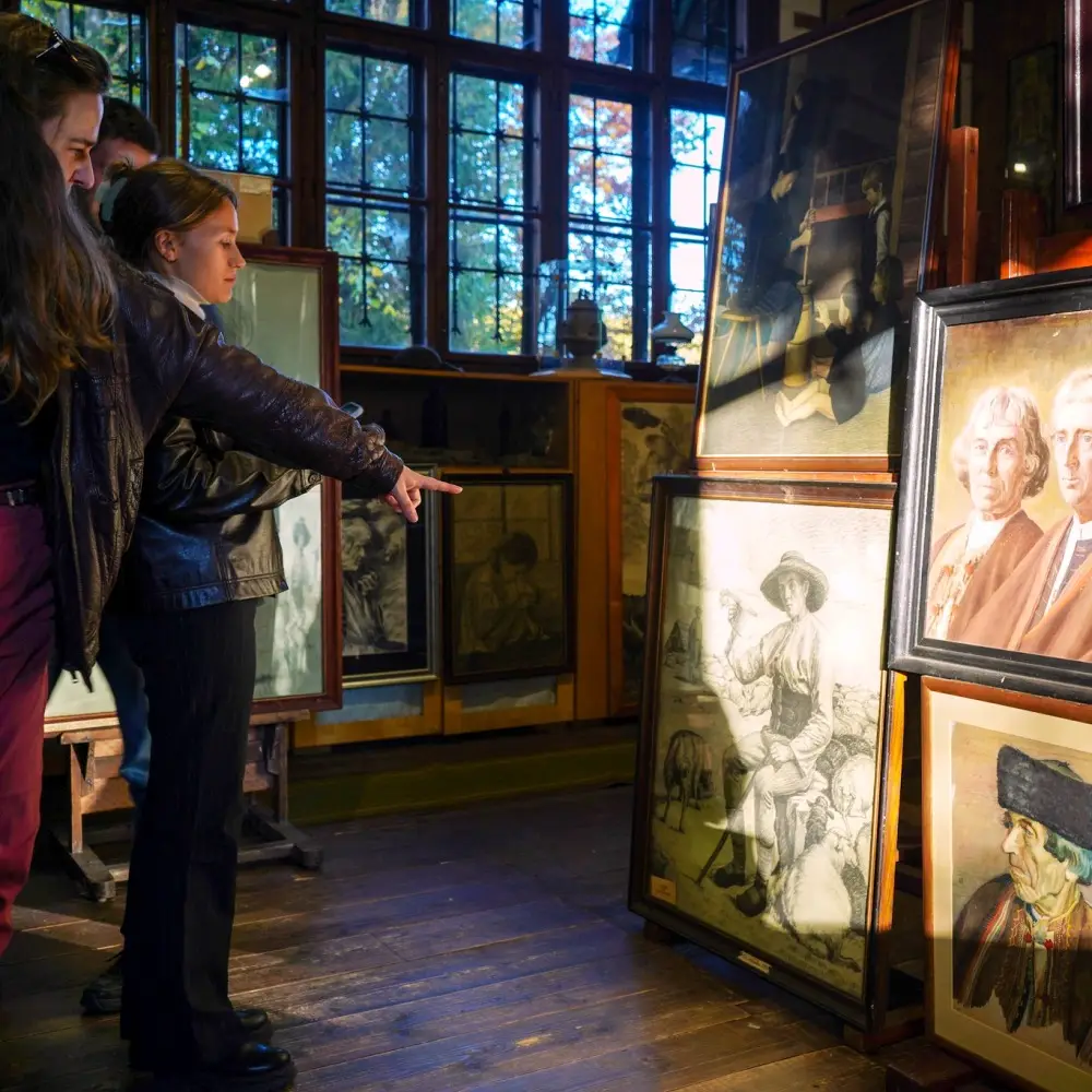 A group of people stands in an art gallery with wooden floors, intently viewing and discussing framed portraits and drawings displayed against a sunlit window. One woman points toward a specific artwork.