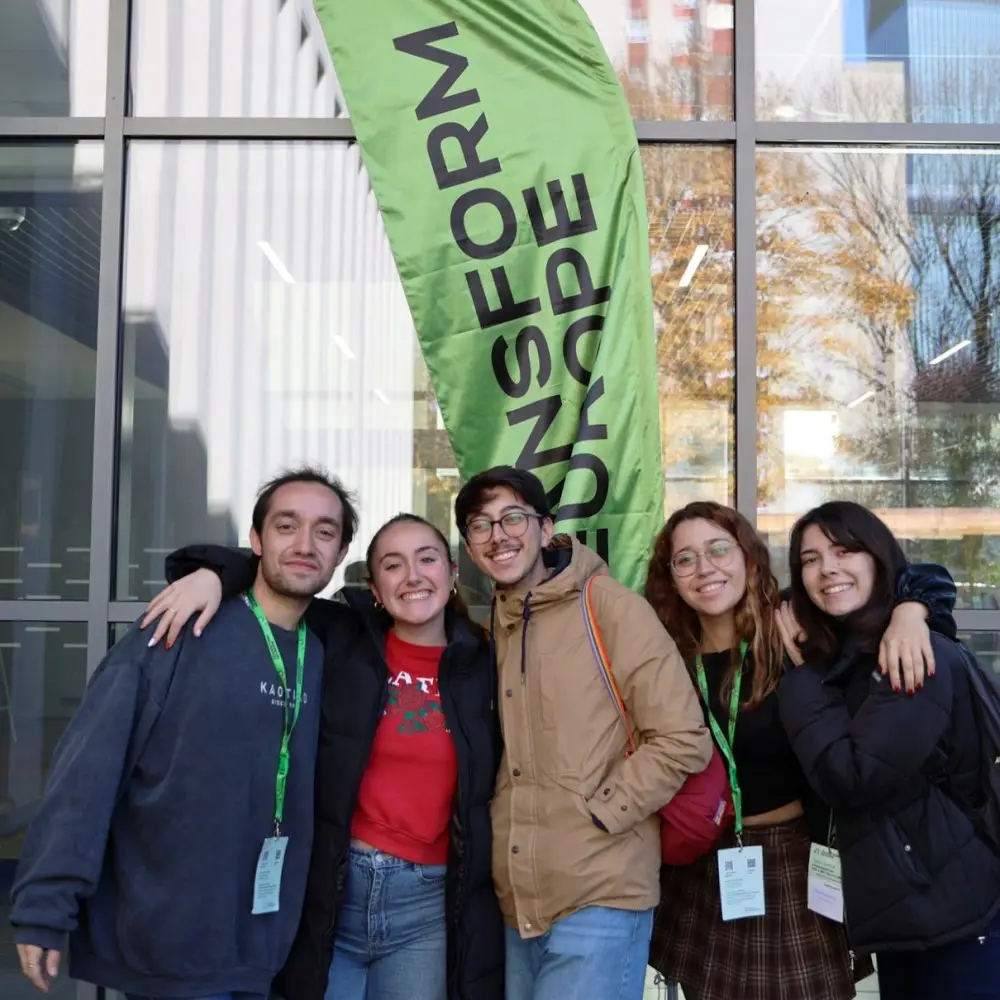 Five young adults stand smiling with arms around each other in front of a green banner that reads TRANSFORM EUROPE, outside a modern building with large windows.
