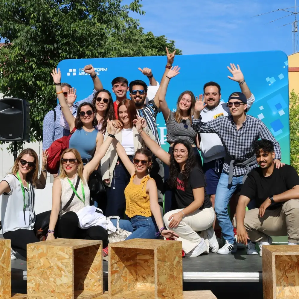 A group of smiling young adults pose together outdoors, some raising their arms and waving, in front of a blue event backdrop on a sunny day. Wooden seats are in the foreground, and trees and buildings are visible behind them.