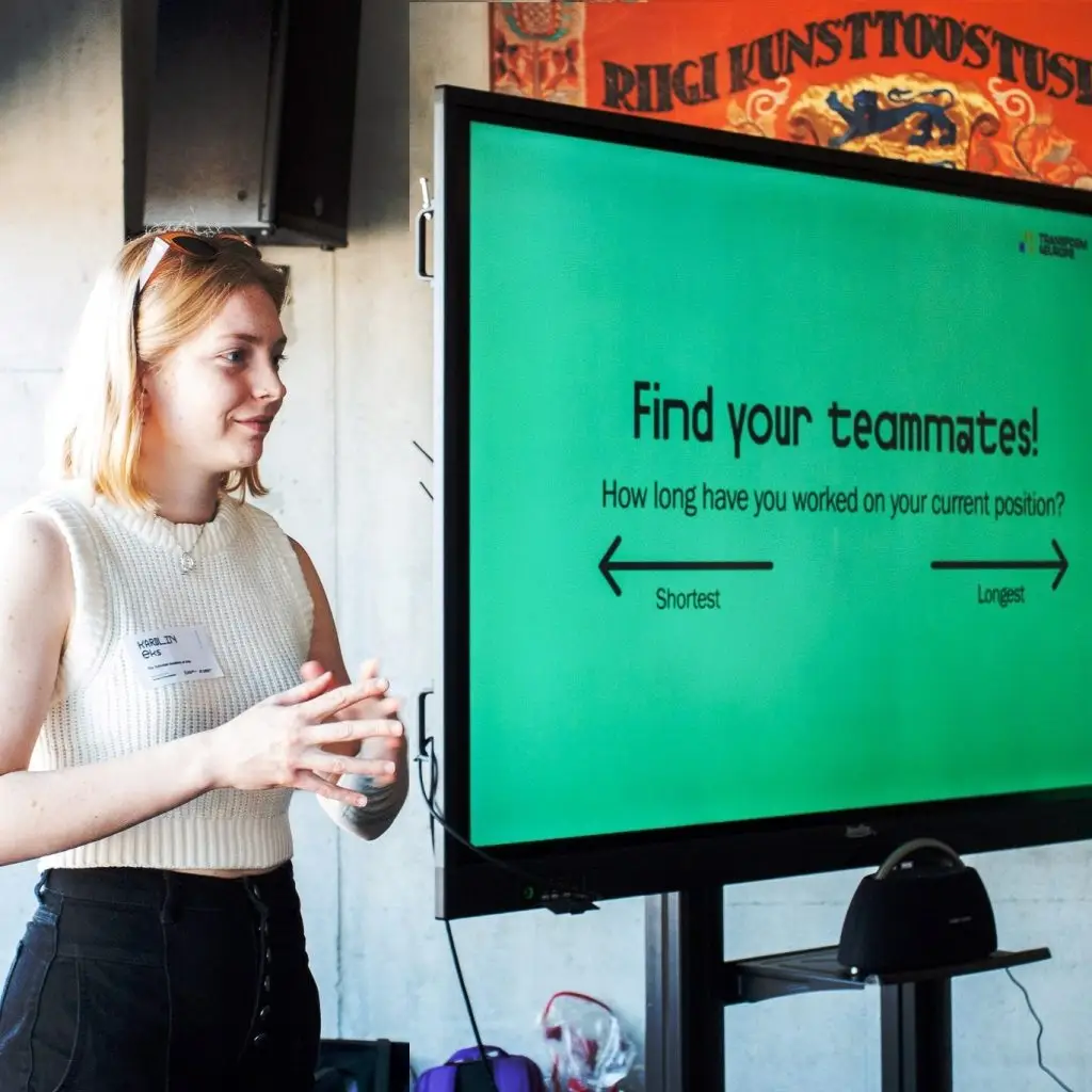 A woman stands beside a large screen displaying the message Find your teammates! How long have you worked on your current position? with arrows pointing to Shortest and Longest. She appears to be presenting.