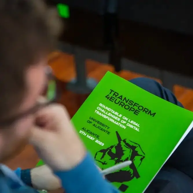 A person holding and reading a bright green booklet titled Transform 4Europe: Roundtable on Legal Challenges of Digital Transformation, with event details for the University of Alicante, 11th May 2023.