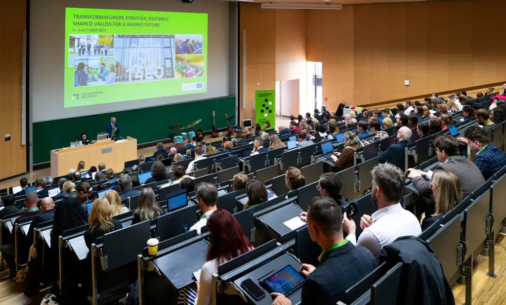 A large lecture hall filled with seated attendees faces a presenter speaking at a podium. A bright presentation slide is projected on a screen at the front, and listeners use laptops and notebooks to take notes.