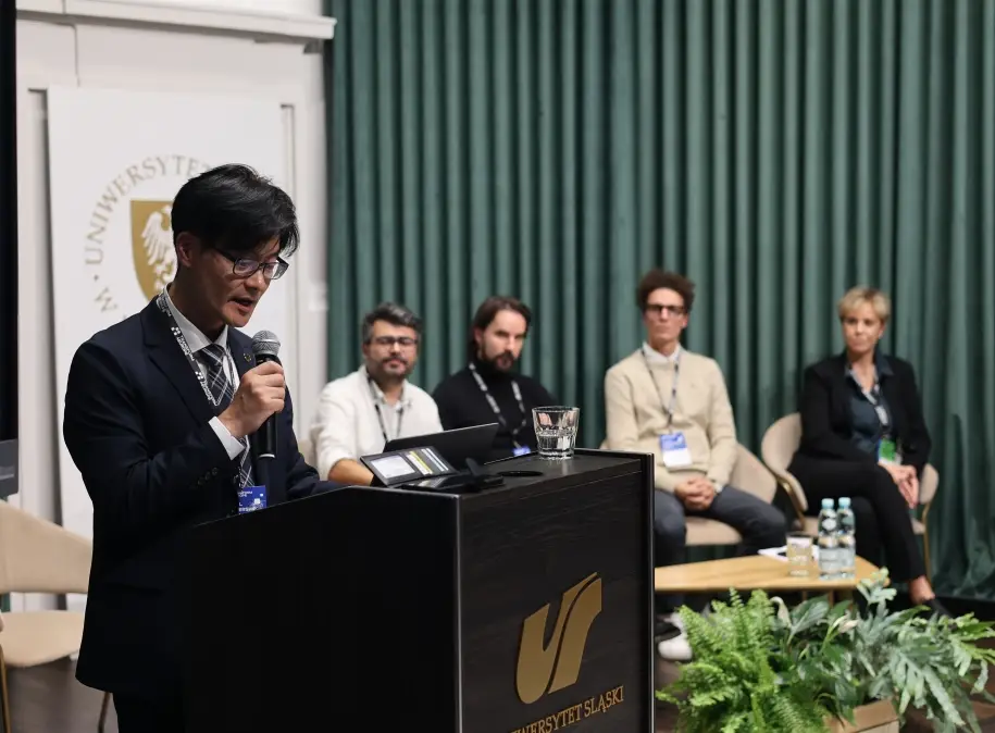 A man speaks at a podium with Uniwersytet Śląski while four people sit behind him on a stage in front of green curtains, suggesting a panel discussion or academic event.