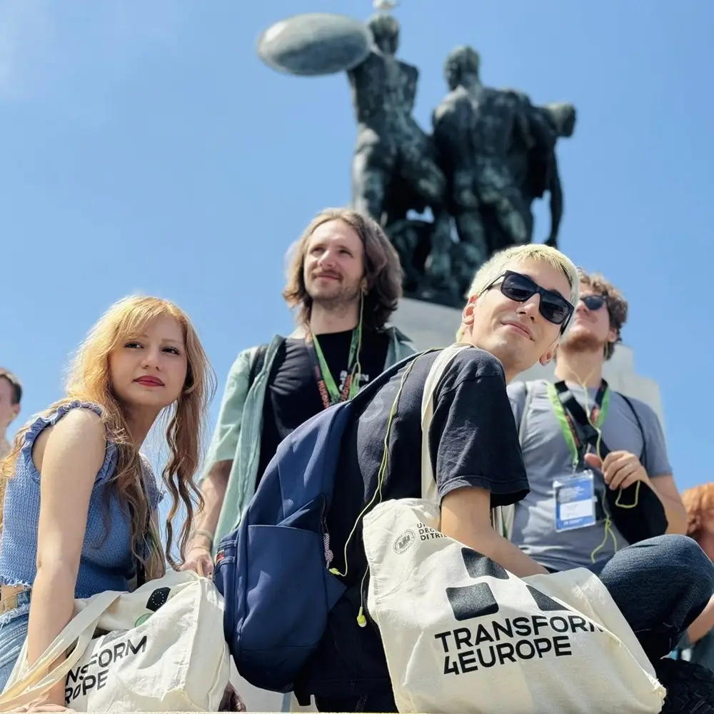 Four young adults with bags labeled Transform4Europe pose in front of a large statue under a sunny sky, looking in different directions. The statue features two figures holding a disc above their heads.