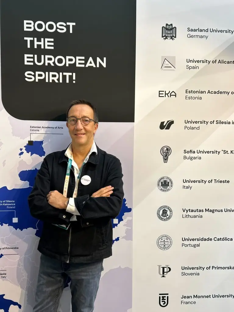 A man in glasses and a dark jacket stands smiling with arms crossed in front of a banner that reads Boost the European Spirit! and lists the names and logos of several European universities.