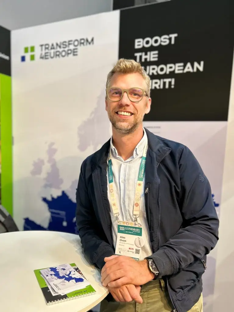 A smiling man with glasses stands at a white table, wearing a conference badge and casual clothes. Behind him are banners with the text TRANSFORM EUROPE and BOOST THE EUROPEAN SPIRIT! and a blue map of Europe.
