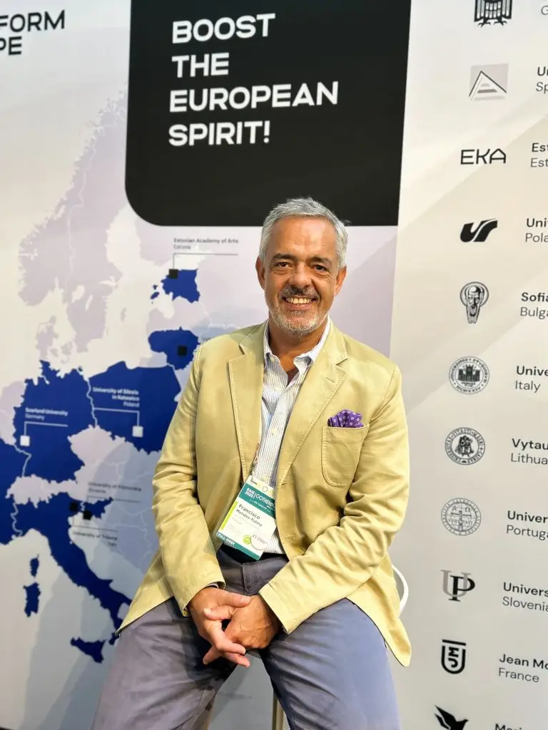 A man in a beige blazer sits and smiles in front of a map of Europe and a sign that reads Boost the European Spirit! Logos of various universities are displayed on the wall behind him.