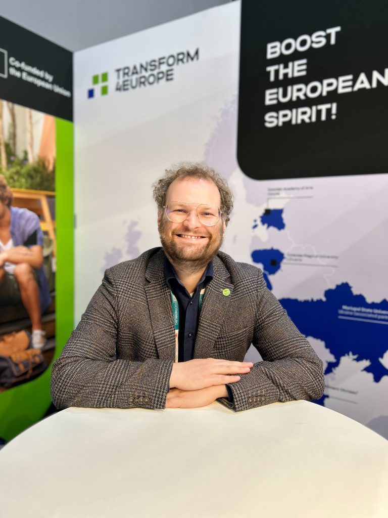 A smiling man with glasses and a beard sits at a table in front of a display reading Transform Europe and Boost the European Spirit! Maps and graphics are visible on the background panels.