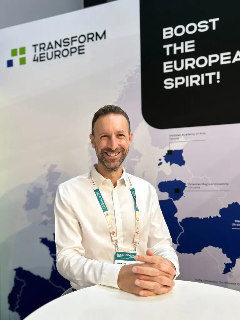 A man in a white shirt sits smiling at a table in front of a display reading TRANSFORM 4EUROPE and BOOST THE EUROPEAN SPIRIT! with a map and university names in the background.