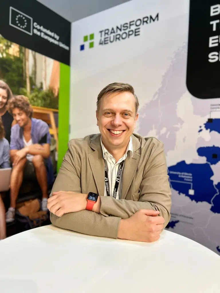 A smiling man in a beige blazer sits at a table with arms crossed. Behind him are banners that read “Transform 4 Europe” and show maps and logos, indicating an academic or professional event.