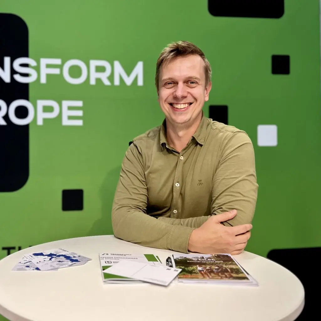 A smiling man in a green shirt sits at a round table with open brochures and papers. Behind him is a green wall with the partial words TRANSFORM EUROPE in white text.