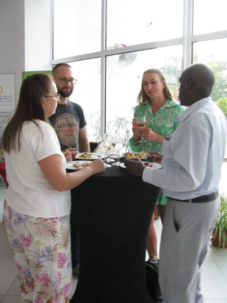 Four adults, two women and two men, stand around a tall round table with drinks and plates of food, socializing in a bright room with large windows and plants in the background.
