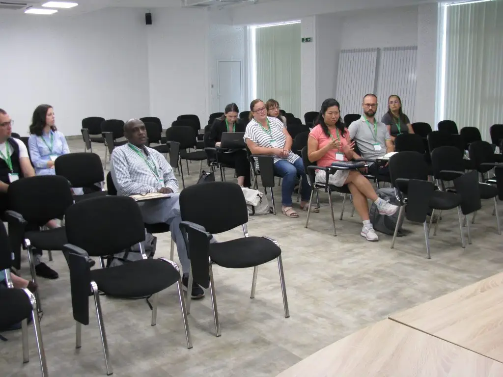 A small group of people sit spaced out on black chairs in a conference room, listening to a woman in a pink shirt and shorts speaking. Some have laptops, and the room has large windows and a neutral color scheme.