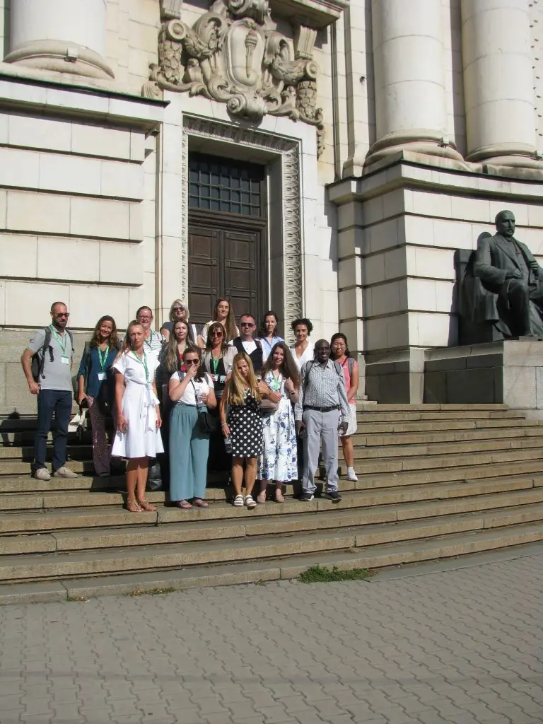A group of people pose for a photo on stone steps outside a large historic building with columns and a statue seated to the right. It’s sunny and everyone is looking at the camera, some smiling.