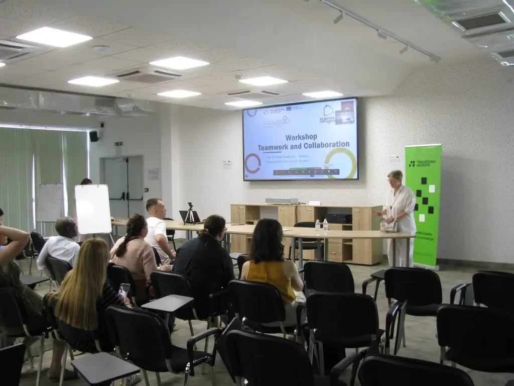 A group of people sit facing a presenter in a classroom, attending a workshop titled Teamwork and Collaboration displayed on a large screen at the front of the room.