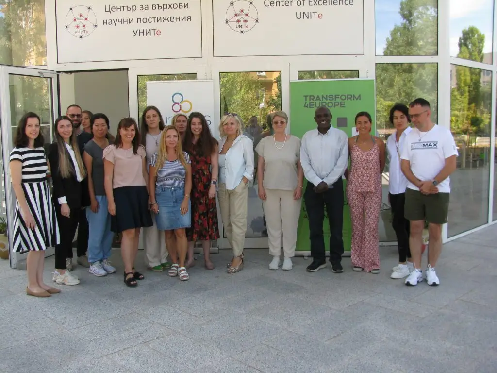 A group of fifteen people, both men and women, stand together in front of glass doors with signs reading Center of Excellence UNITE and TRANSFORM 4EUROPE. They are posing and smiling at the camera outside.