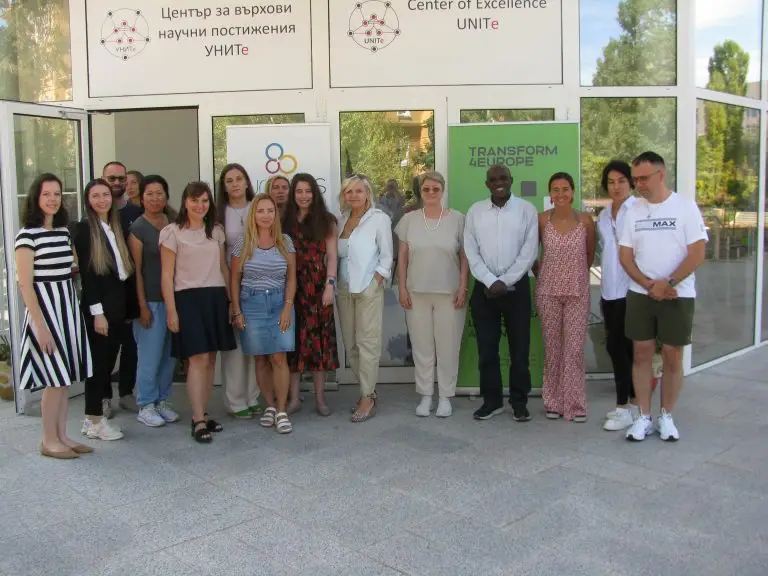 A group of fifteen people, both men and women, stand together in front of glass doors with signs reading Center of Excellence UNITE and TRANSFORM 4EUROPE. They are posing and smiling at the camera outside.