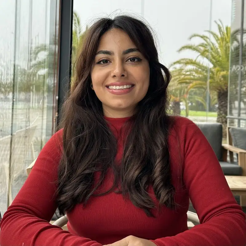 A woman with long dark hair and a red top sits at a table in a cafe, smiling at the camera. Palm trees and large windows are visible in the background on a cloudy day, setting the scene for a Transform for Europe gathering.