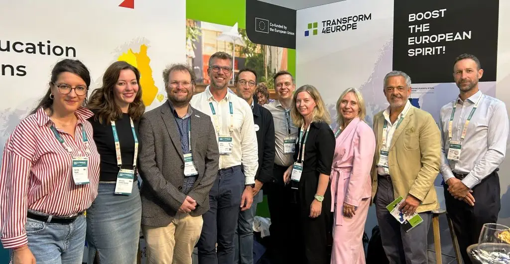 A group of ten people stand together and smile at a conference booth with banners reading “Transform 4 Europe” and “Boost the European Spirit!” Some wear lanyards and business attire.