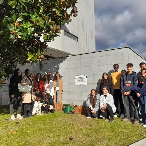 A group of 17 young people stands and kneels together on green grass beside a building under a large tree, posing and smiling in the sunlight near a sign on the wall.