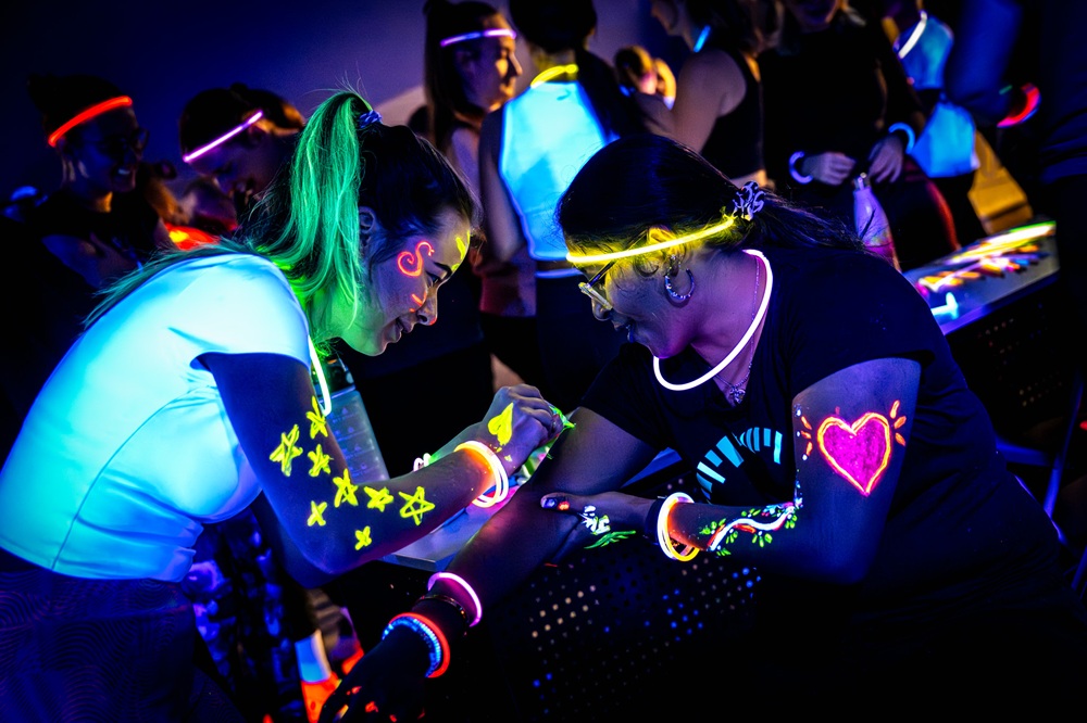People at a glow-in-the-dark party wear neon face paint and glow sticks under black lights. Two women are focused as one paints glowing designs on the others arm. The scene is lively with vibrant, fluorescent colors.
