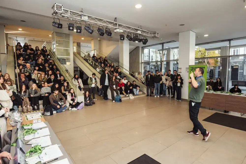 A man speaks to a large audience seated and standing in a modern indoor space, with stairs and windows in the background. The crowd listens attentively, and event tables with papers are set up on the left.