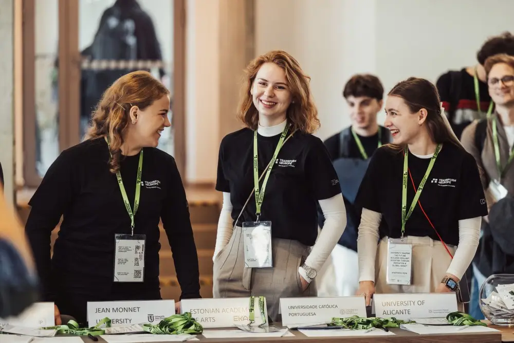 Three young women wearing black shirts and name badges stand behind a table with university name cards, smiling and talking at an indoor event. Other people are visible in the blurred background.