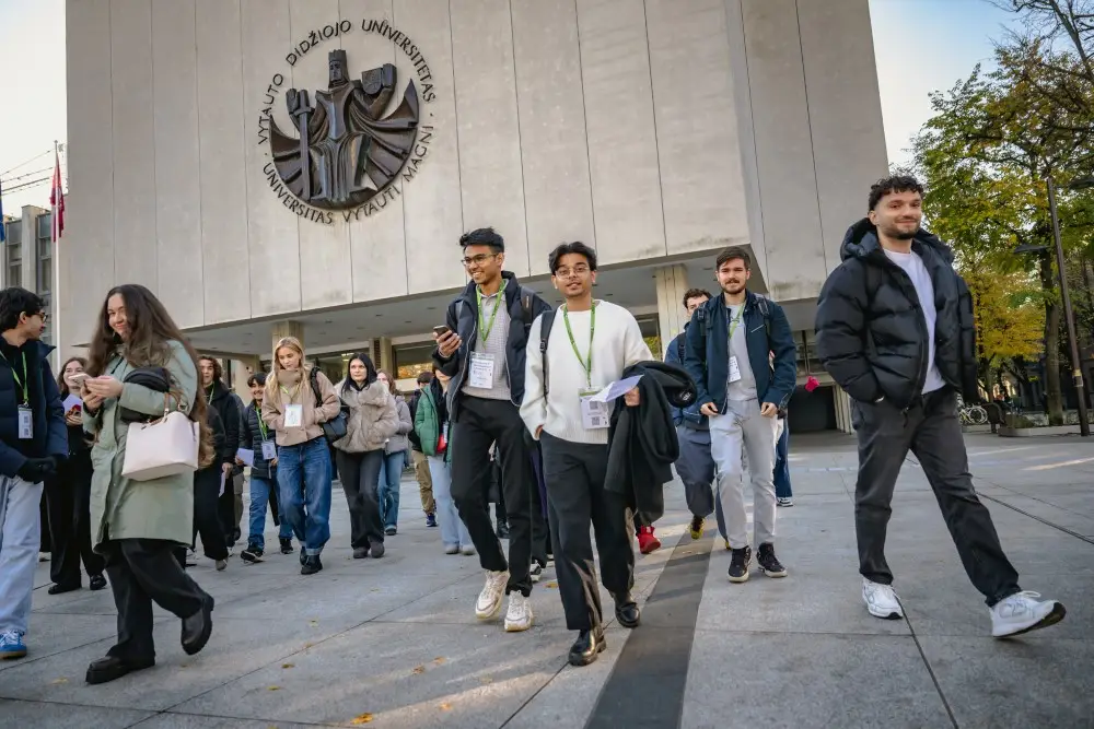 A group of young adults walks out of a university building, smiling and chatting. The entrance features a large emblem and “Vytauto Didžiojo Universitetas” written above the doors. Some people wear lanyards and carry papers.