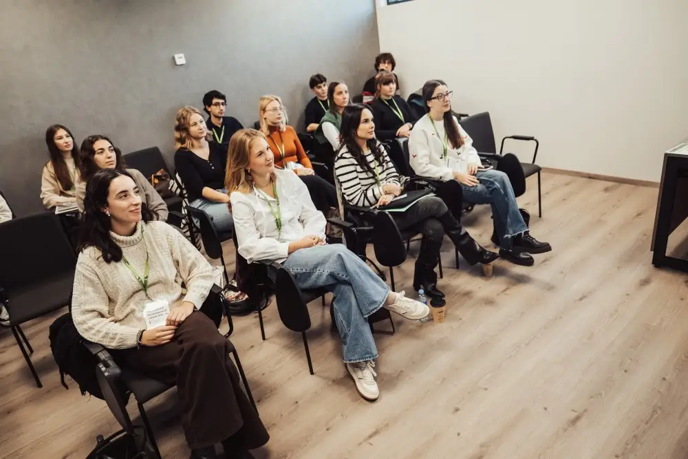 A group of young adults sits in rows of chairs in a modern classroom, attentively listening to a presentation. Most have notepads or laptops, and some wear lanyards. The room has light wood floors and gray walls.