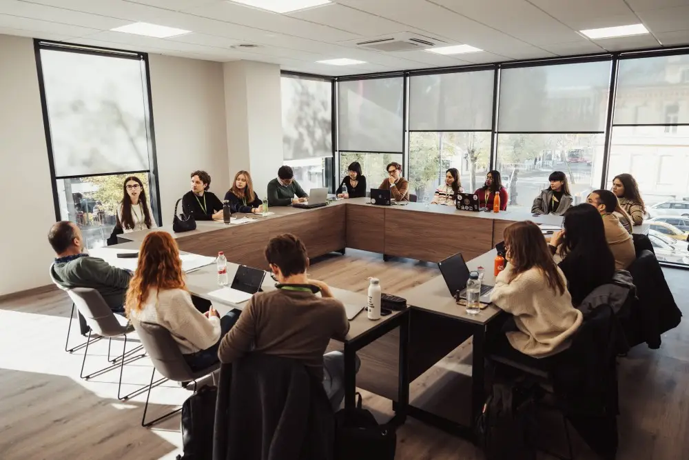 A group of people sits around a large U-shaped conference table in a bright, modern meeting room with large windows. Some have laptops open and everyone appears to be engaged in discussion.