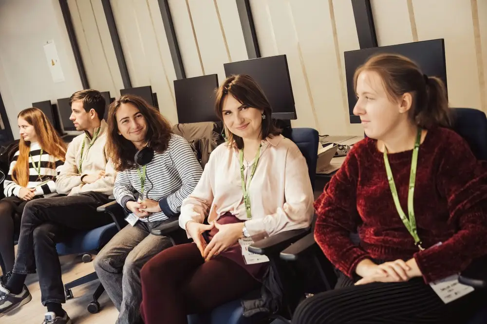 A group of five young adults with lanyards sit in a row of office chairs in front of computer monitors, smiling and conversing in a modern classroom or workspace.