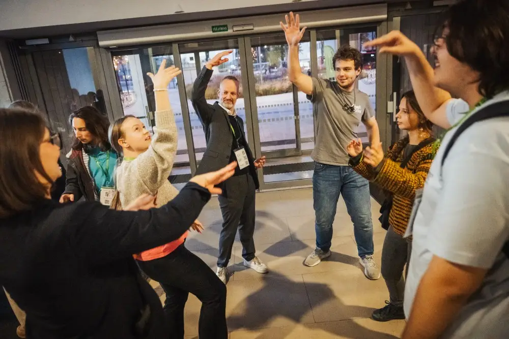 A group of people stand in a circle indoors, smiling and raising their arms enthusiastically, as if playing an interactive game or engaging in an energetic group activity.