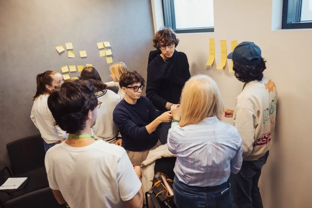 A group of people stands in a small room, collaborating and placing sticky notes on the walls. Some are writing or discussing ideas, while others observe and participate in the brainstorming session.