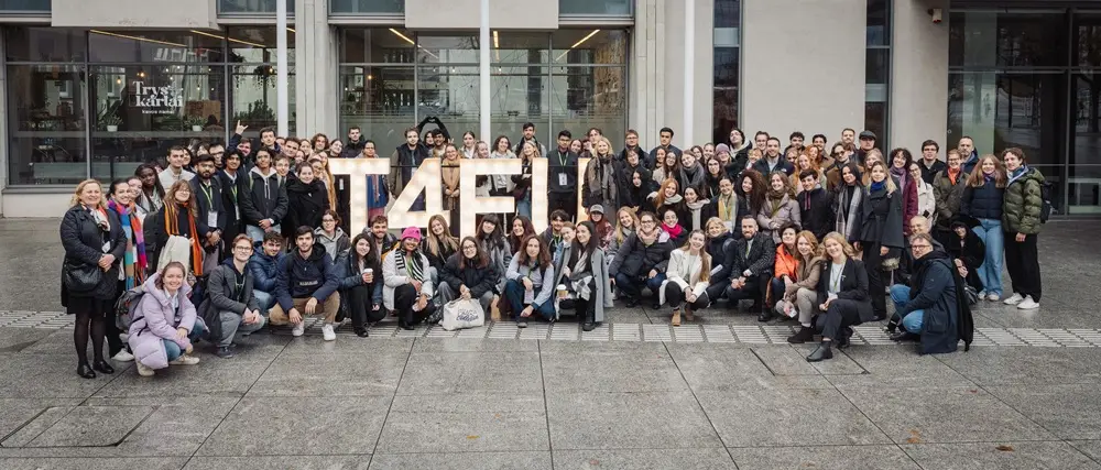 A large group of people pose together outdoors in front of a building, gathered around large, illuminated letters spelling T4EU. Most are smiling and dressed in jackets and winter clothing.