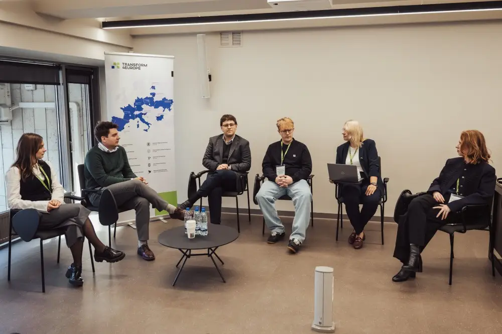 Six people sit in a semicircle on chairs in a modern conference room, engaged in discussion. Behind them is a banner with a map of Europe, and a round table with water bottles is in the center.